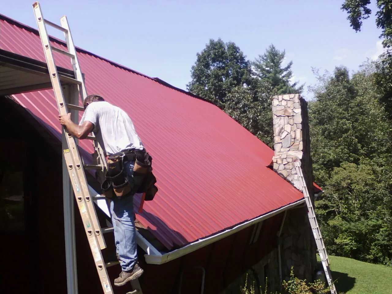 Roofer on a ladder beside a red metal roof and stone chimney — Soffit & Fascia Repair by Roofing Brothers in Strafford